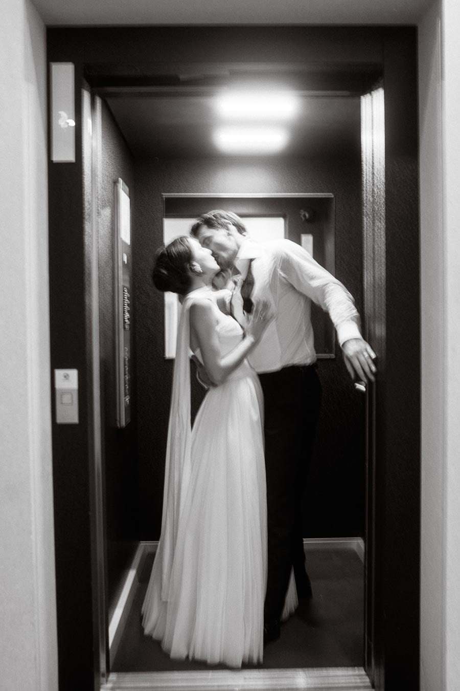 A romantic black and white photo of a couple sharing a passionate kiss in an elevator, with the woman wearing an elegant flowing dress and the man in a shirt and tie, capturing an intimate and timeless moment.