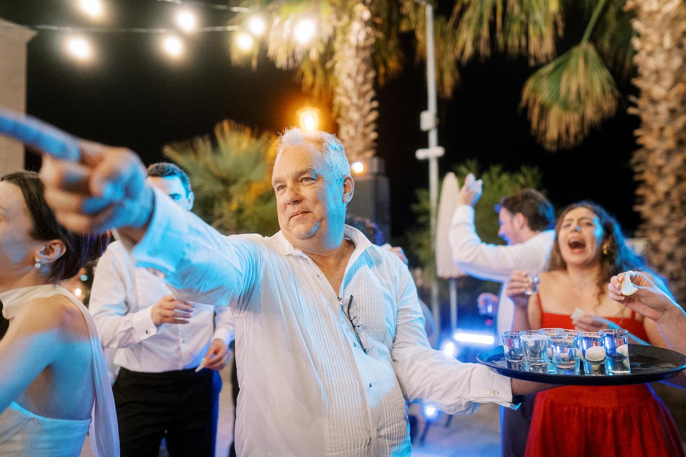 A lively outdoor party scene with a man in a white shirt holding a tray of shot glasses, surrounded by joyful guests under palm trees and string lights at night.