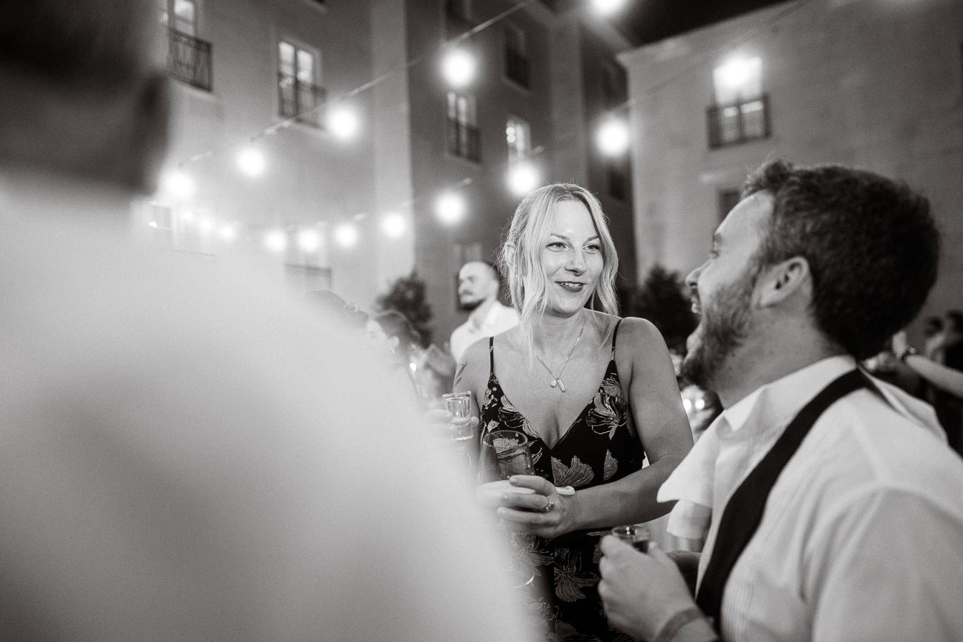 Black and white photo of a woman in a floral dress smiling and talking to a man during an outdoor evening gathering, with string lights illuminating the background.