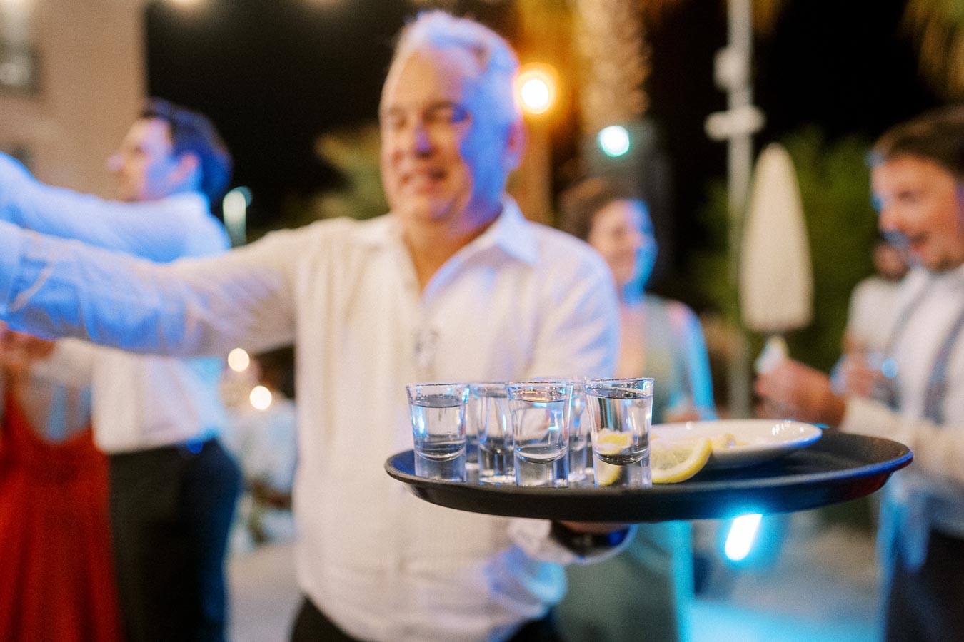 Man serving shots of alcohol on a tray at a lively outdoor evening event, with blurred background of people celebrating and colorful lights.