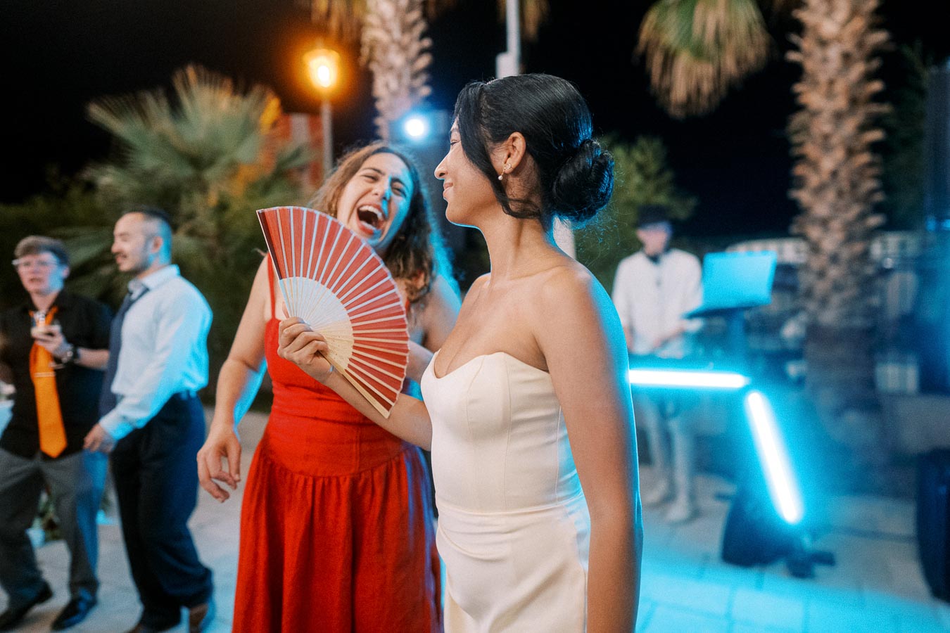 Women laughing and enjoying a night-time outdoor party, with one in a white dress holding a red fan. The scene is lively with colorful lights and palm trees in the background, creating a festive atmosphere.