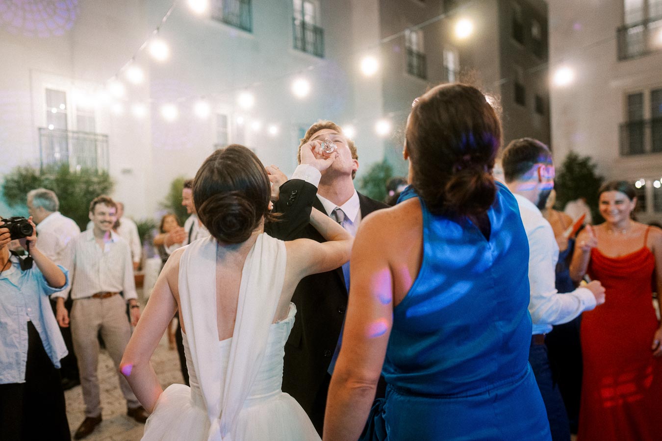 A lively wedding reception scene with guests celebrating outdoors under string lights. A person in a white dress and another in a blue dress share a drink with a suited man, surrounded by smiling friends.