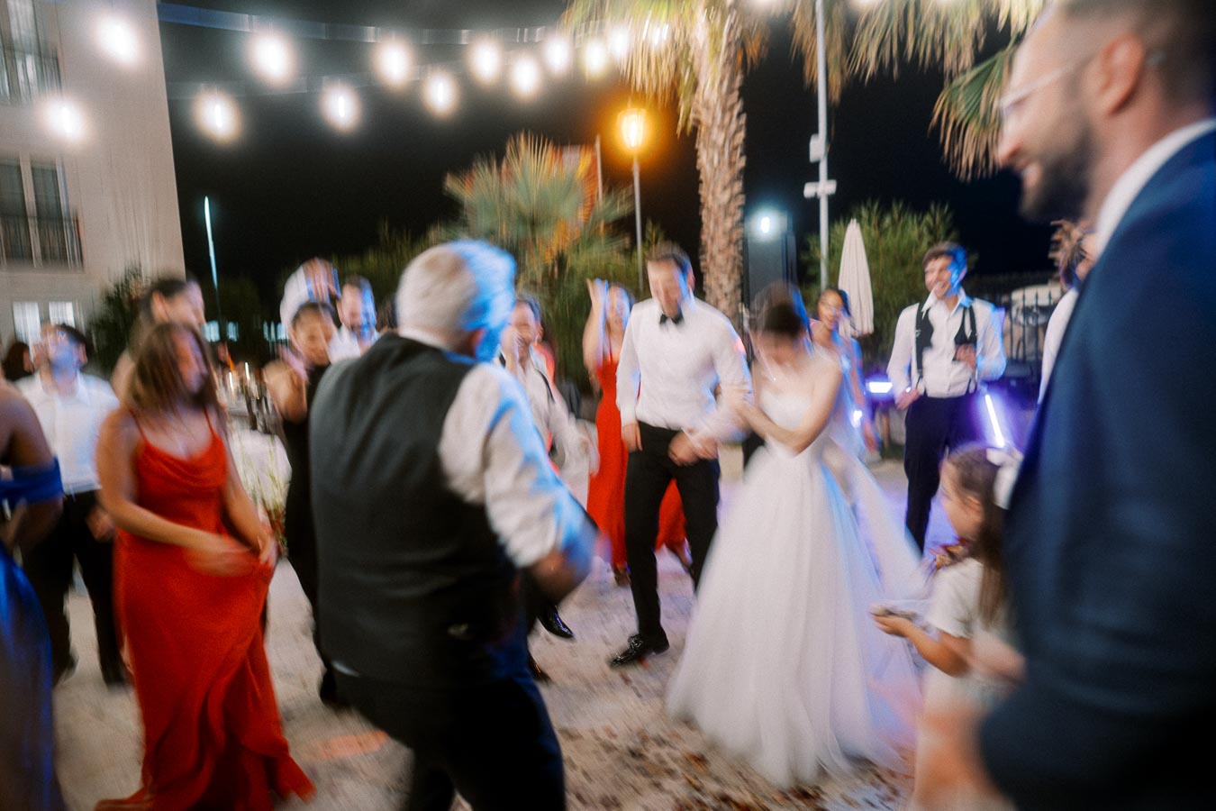Blurry wedding reception scene with guests dancing energetically outdoors under string lights, including a bride in a white gown and others dressed in formal attire, surrounded by palm trees.