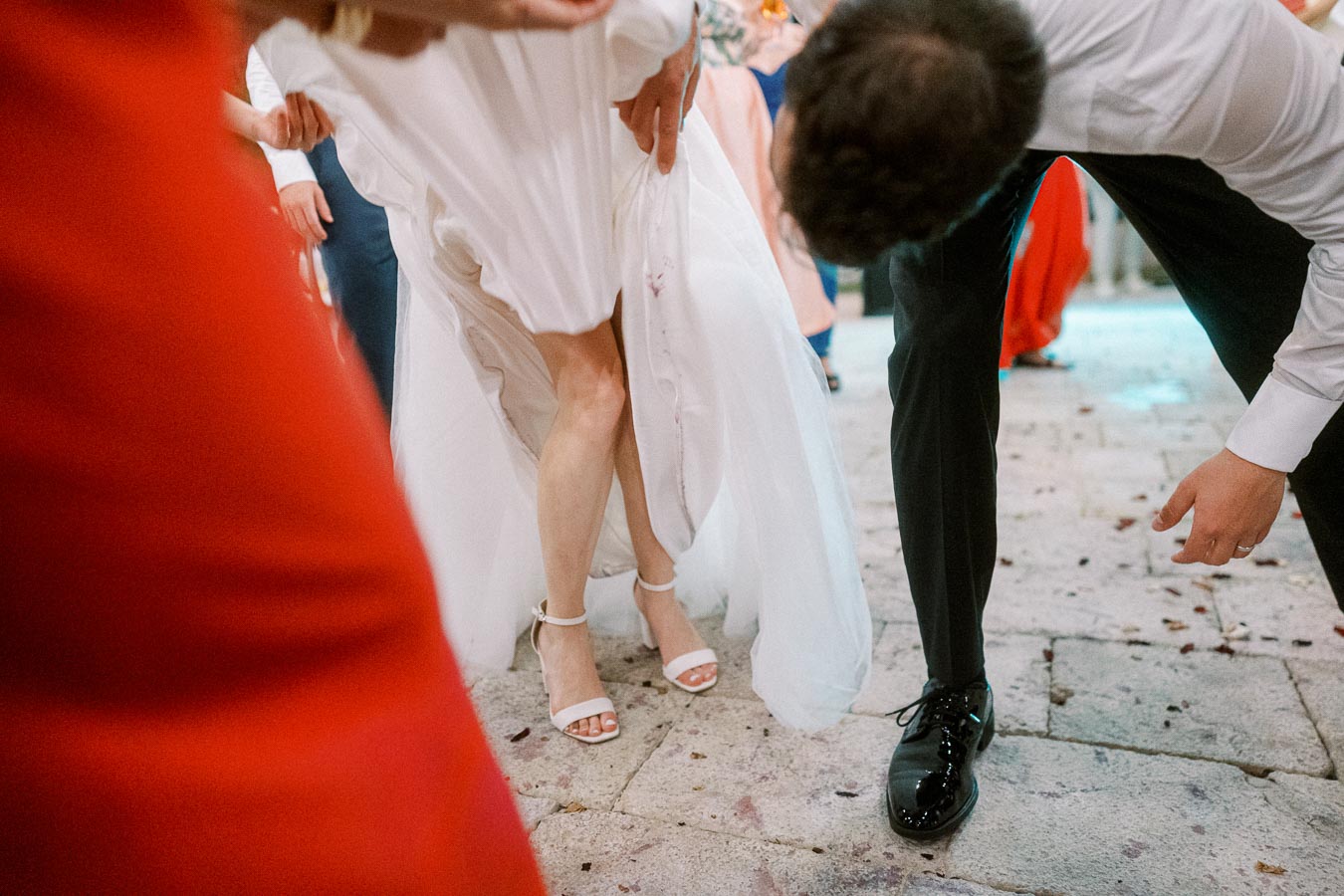 Wedding guests dancing energetically on a stone floor, with a bride in a white dress and white heels lifting her skirt slightly, surrounded by people in formal attire.