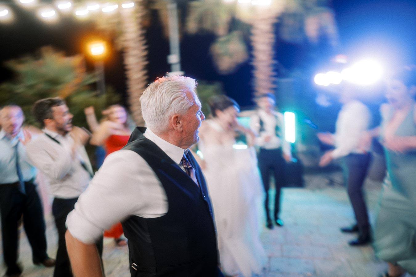 A lively wedding reception with guests dancing under outdoor string lights, featuring a gray-haired man in a black vest in the foreground.