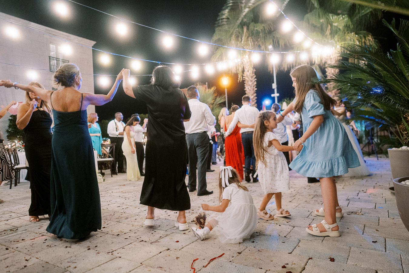 Outdoor evening wedding celebration with guests dancing under string lights, featuring children playing and vibrant dresses, palm trees in the background.