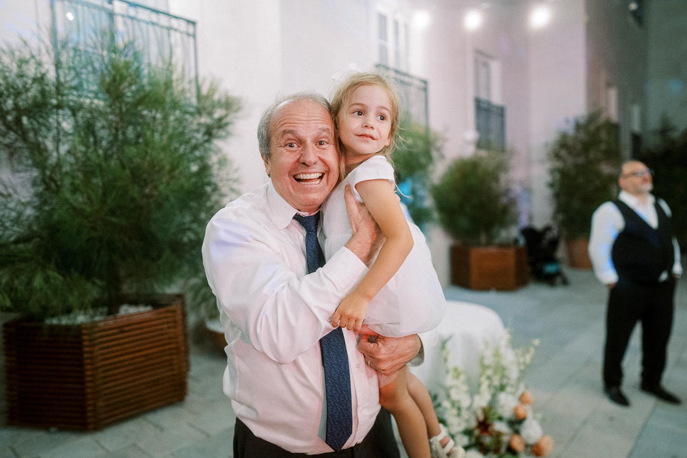 Elderly man joyfully holds a young girl in his arms at an outdoor event, both smiling, surrounded by greenery and soft lighting.