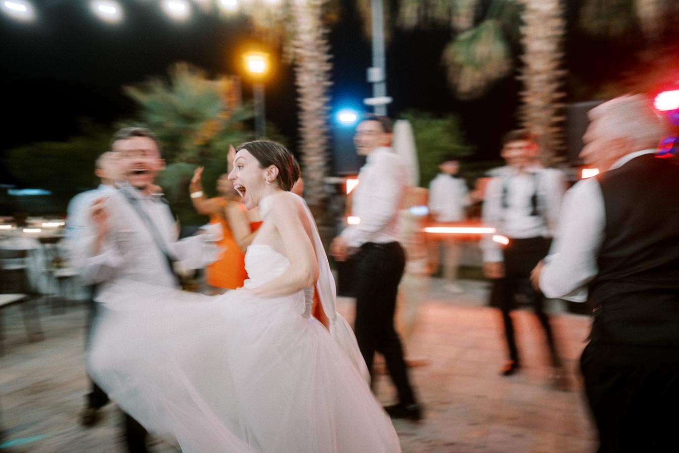 Joyful bride dancing with guests at an outdoor night wedding reception under string lights.
