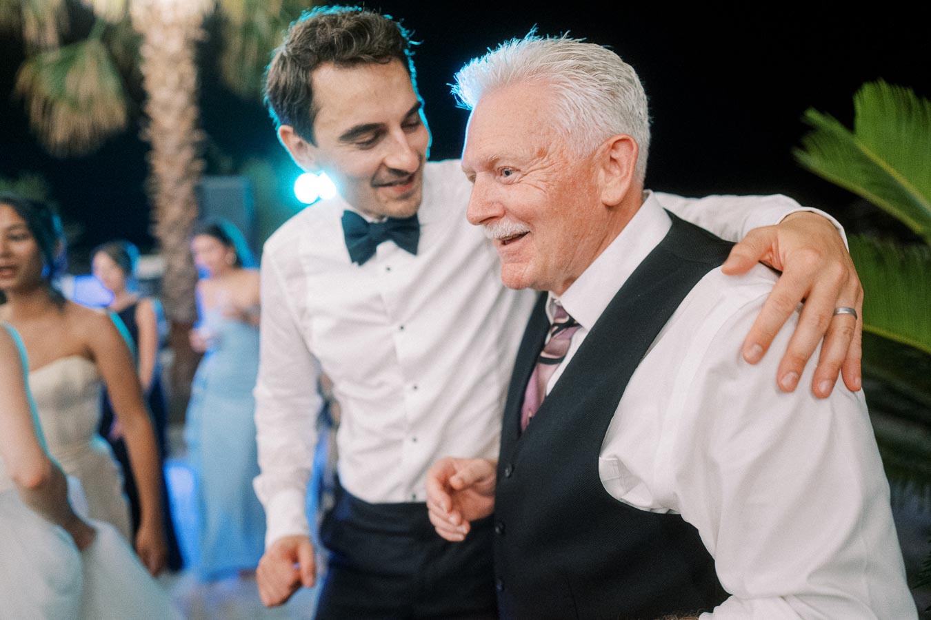 Joyful wedding celebration with two men embracing, surrounded by dancing guests dressed in formal attire.
