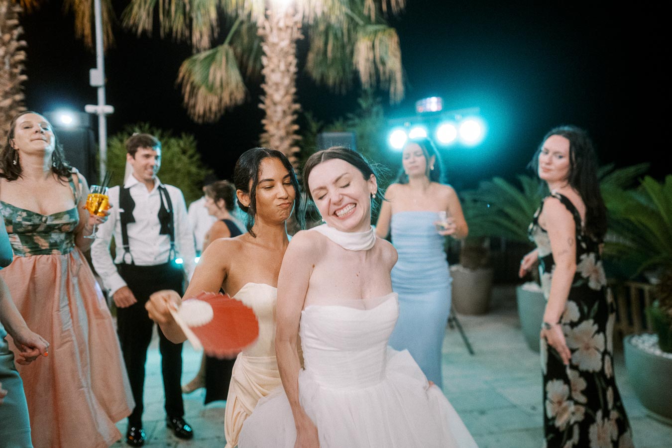 A joyful bride dancing with friends at an outdoor evening wedding reception, with palm trees and bright lights in the background.