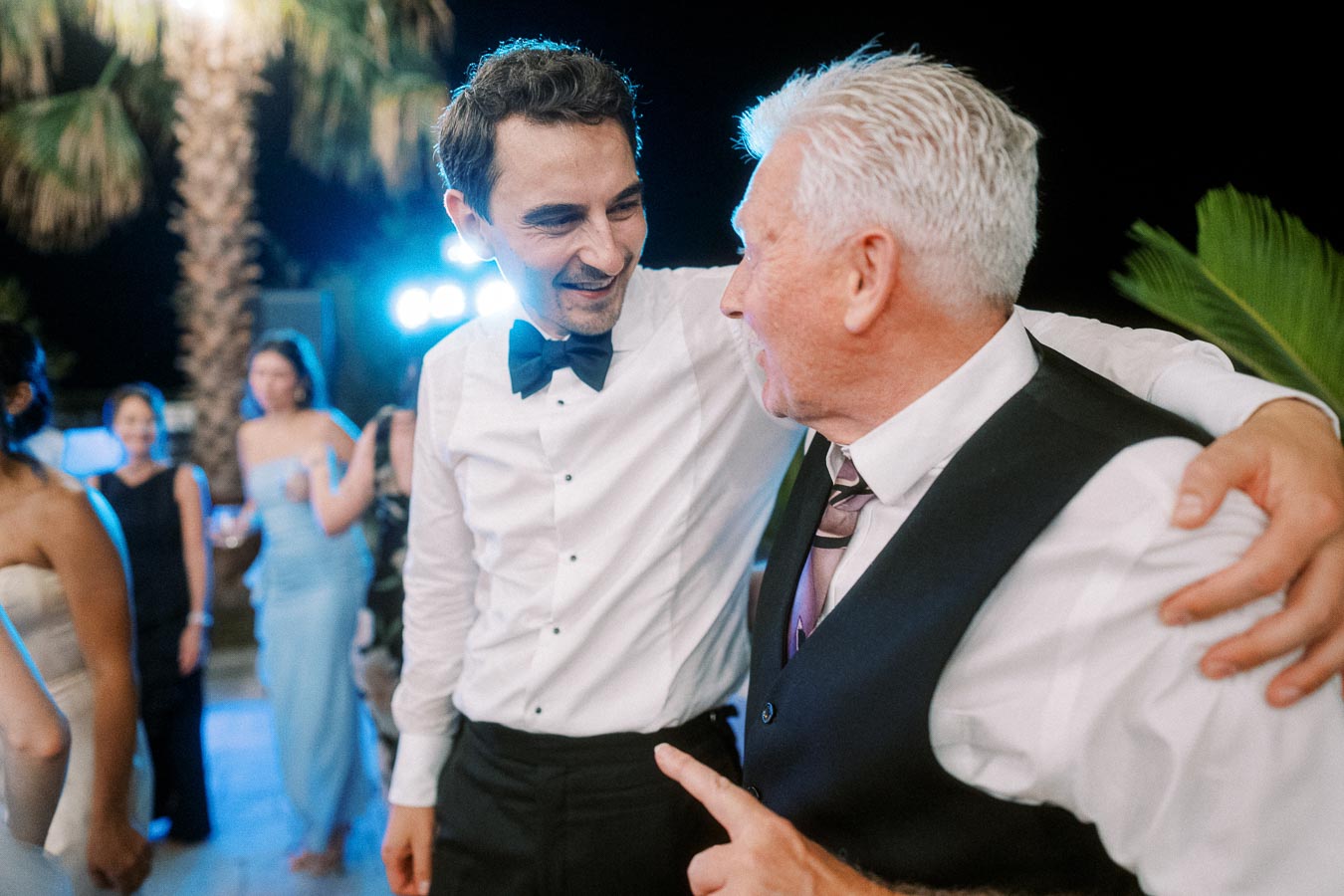 Two men in formal attire sharing a joyful moment at an outdoor evening event with blurred guests in the background.
