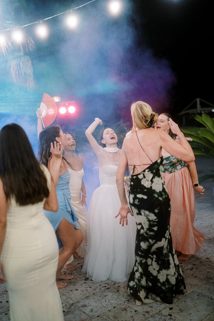 A group of women joyfully dancing at a nighttime wedding reception, surrounded by colorful lights and smoke effects, creating a lively and festive atmosphere.