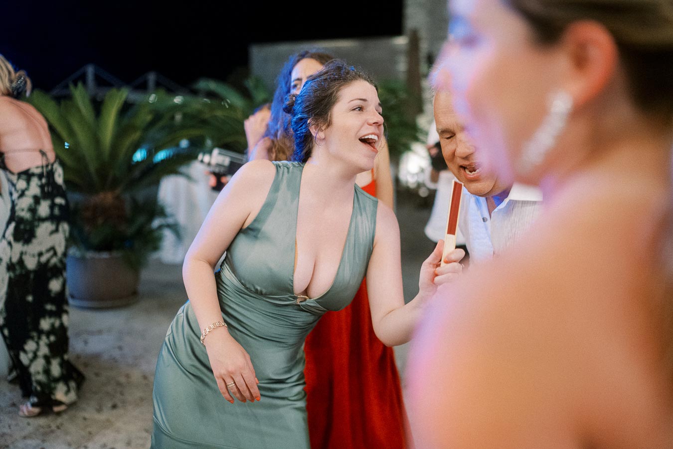 A group of people enjoying a lively outdoor party, with a woman in a teal dress laughing and holding a popsicle, surrounded by others in vibrant attire, against a backdrop of greenery.