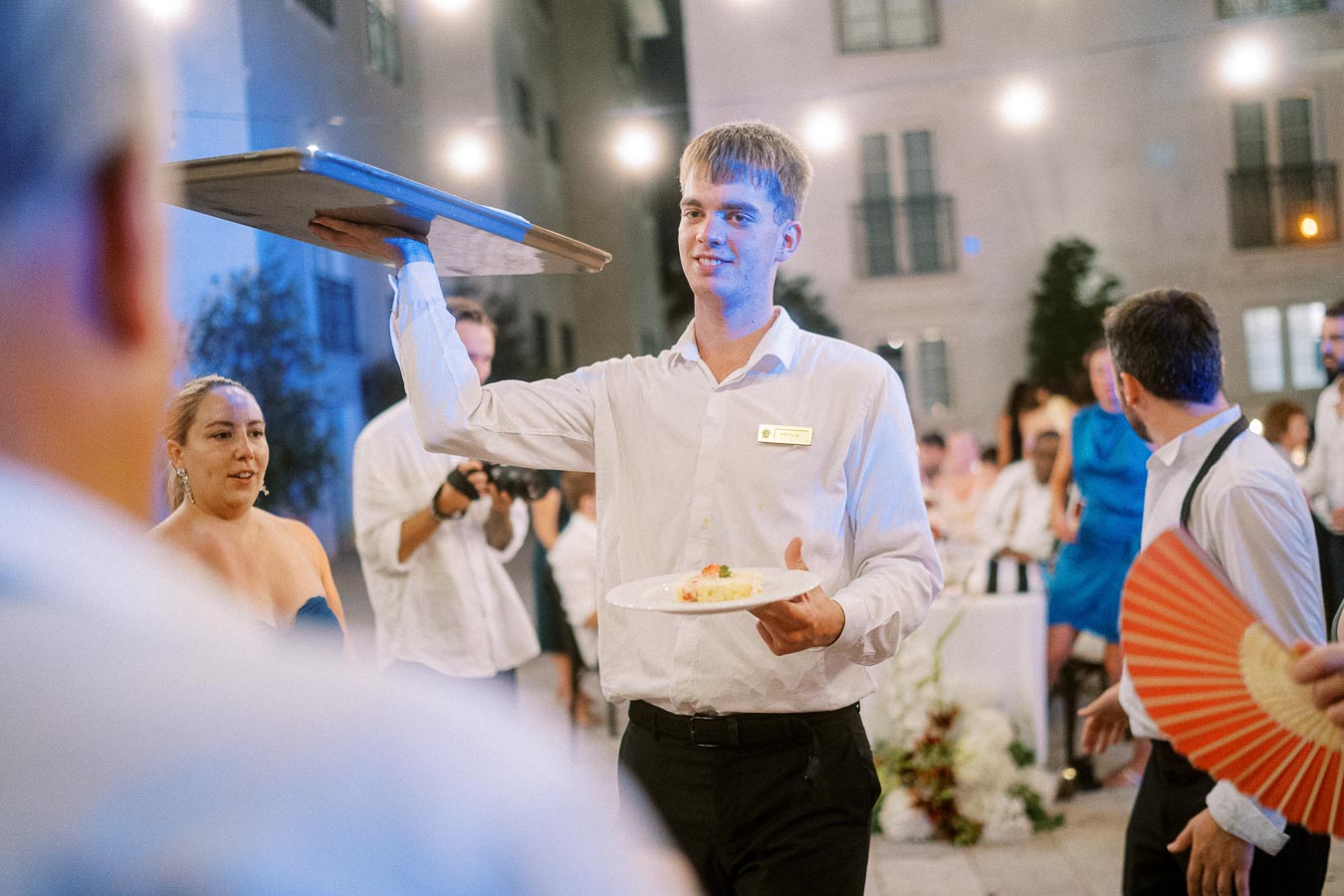 A waiter in a white shirt elegantly balancing a tray and serving food at an outdoor evening event, with guests and lights in the background.