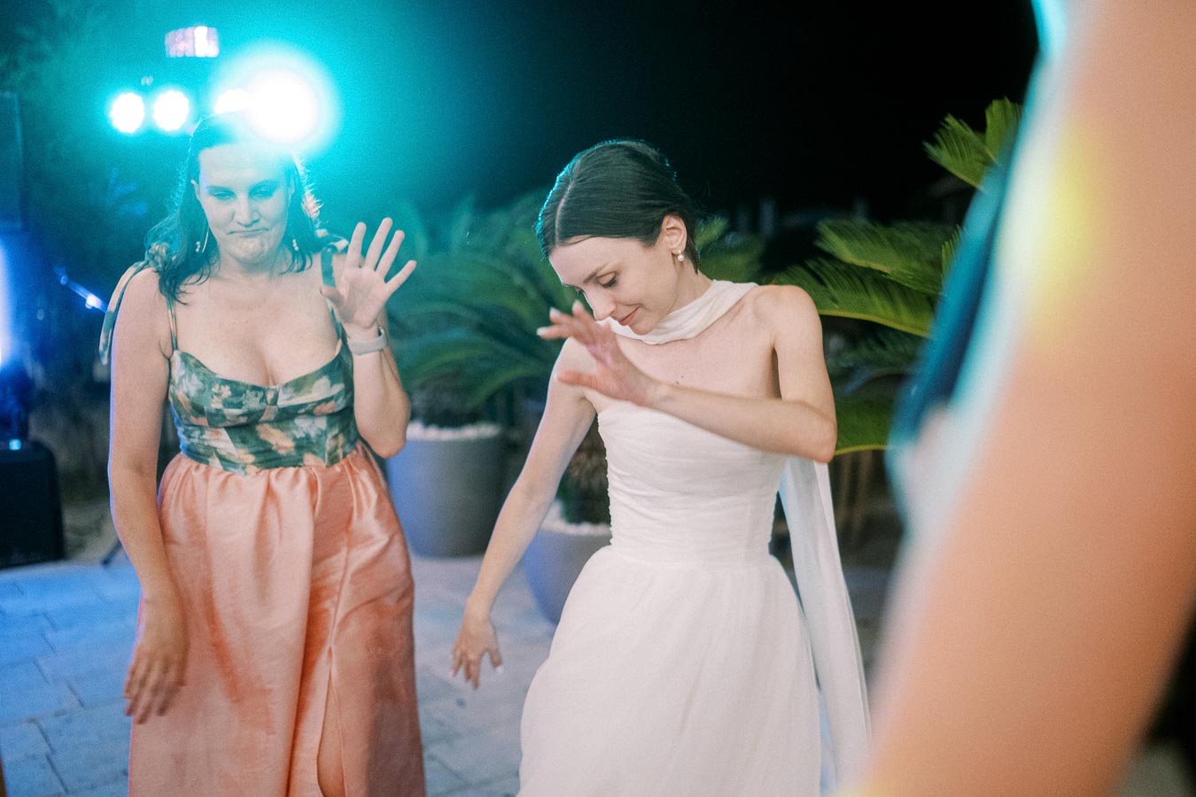 Two women dancing joyfully at an outdoor evening event, illuminated by colorful stage lights, with one in a white dress and the other in a floral top with a pink skirt.