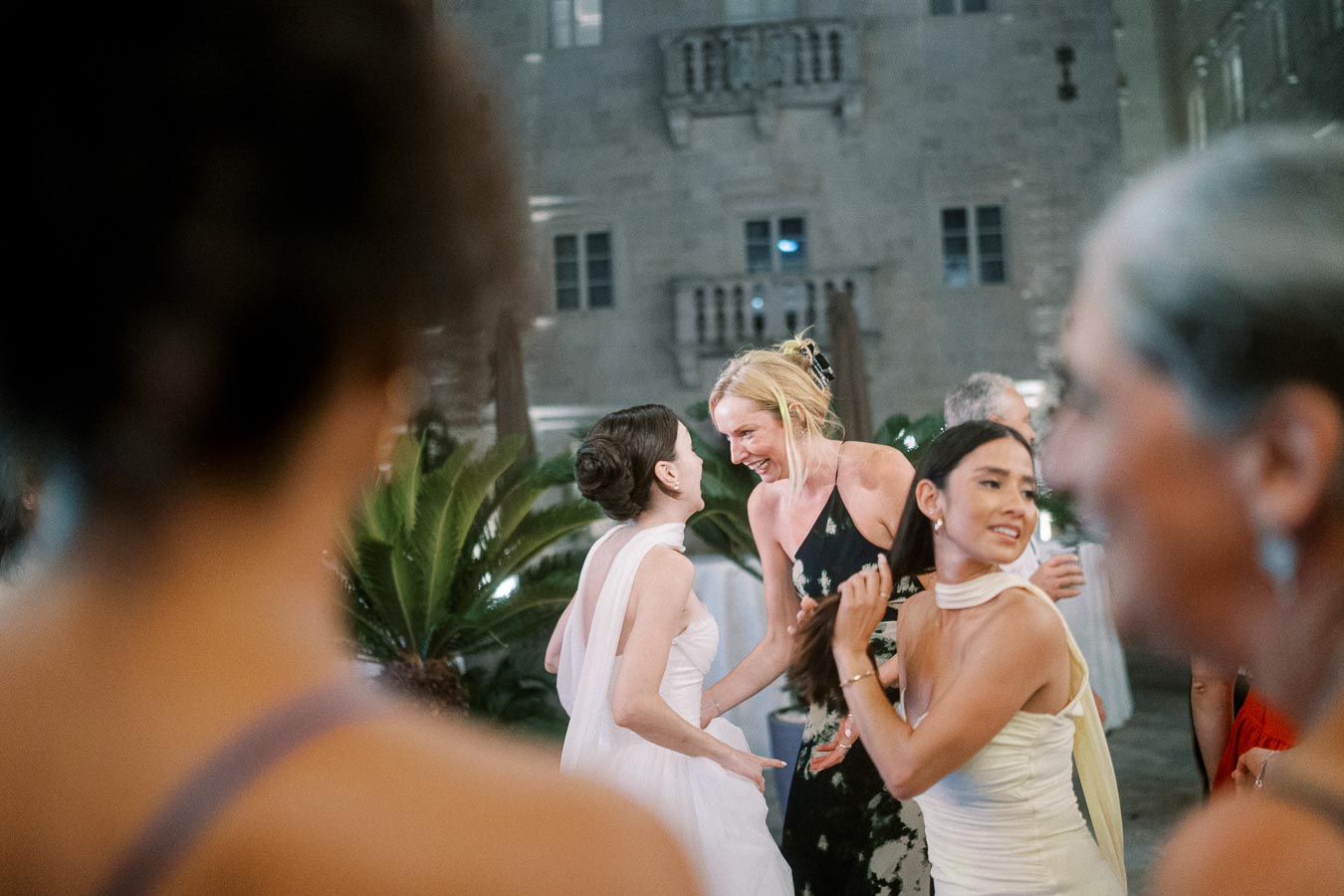 A group of elegantly dressed women engaged in conversation and laughter at an outdoor evening event, with a historic building backdrop and lush greenery.
