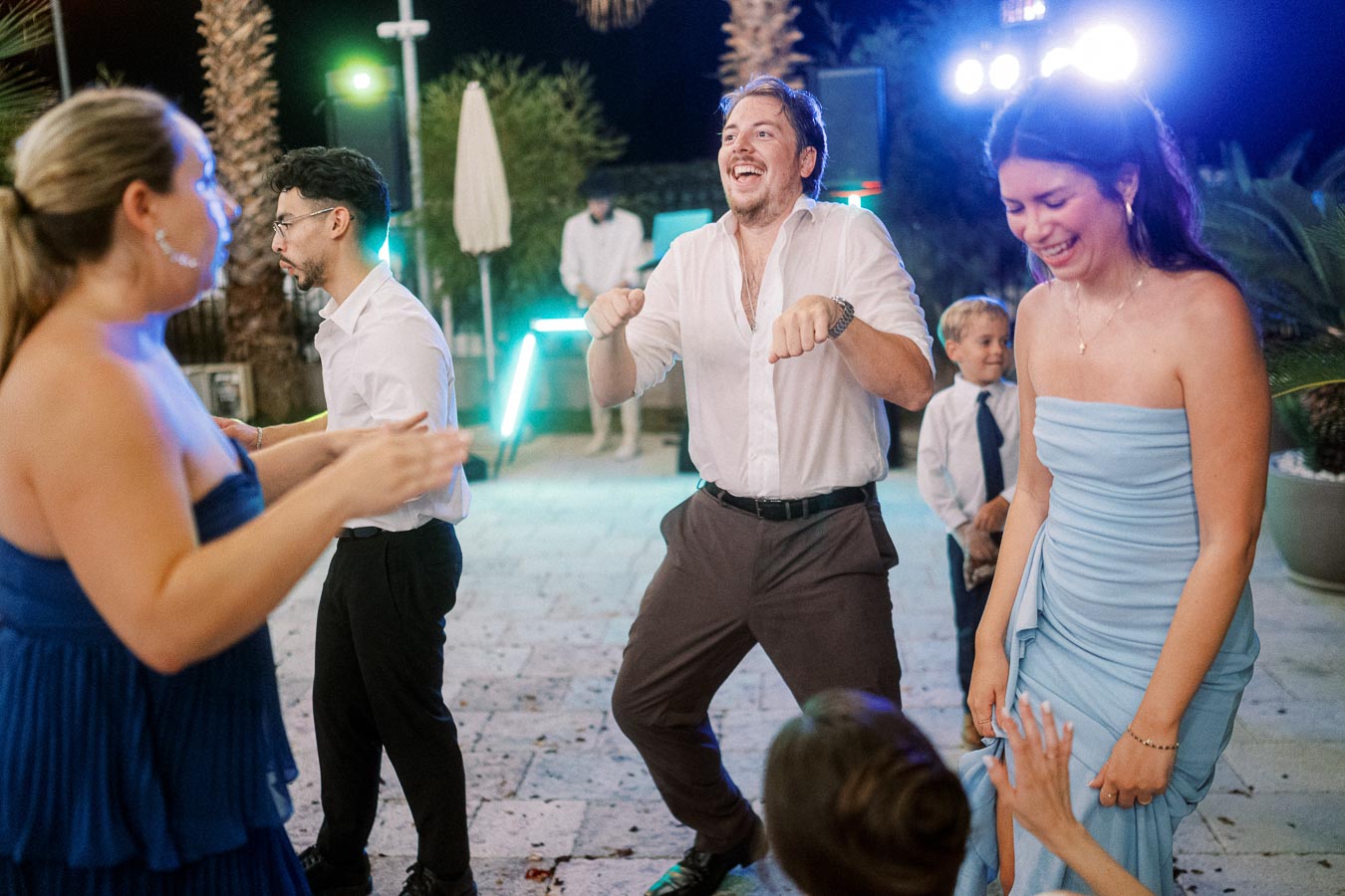 Group of people joyfully dancing at an outdoor evening event, with colorful lighting and palm trees in the background.