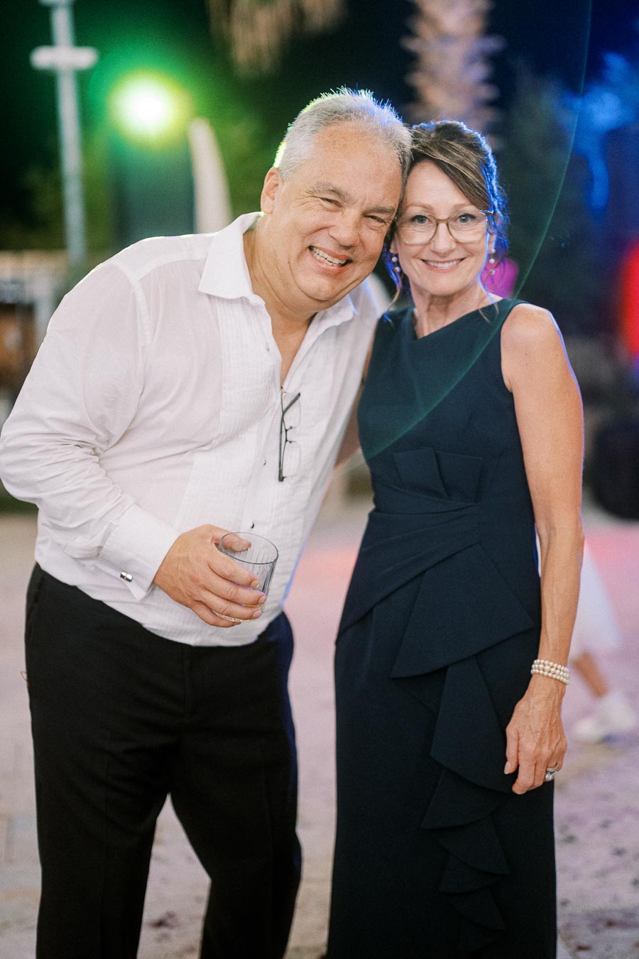 A smiling couple attending an outdoor evening event, the man in a white shirt holding a drink, and the woman in a stylish dark dress with glasses, under vibrant lighting.
