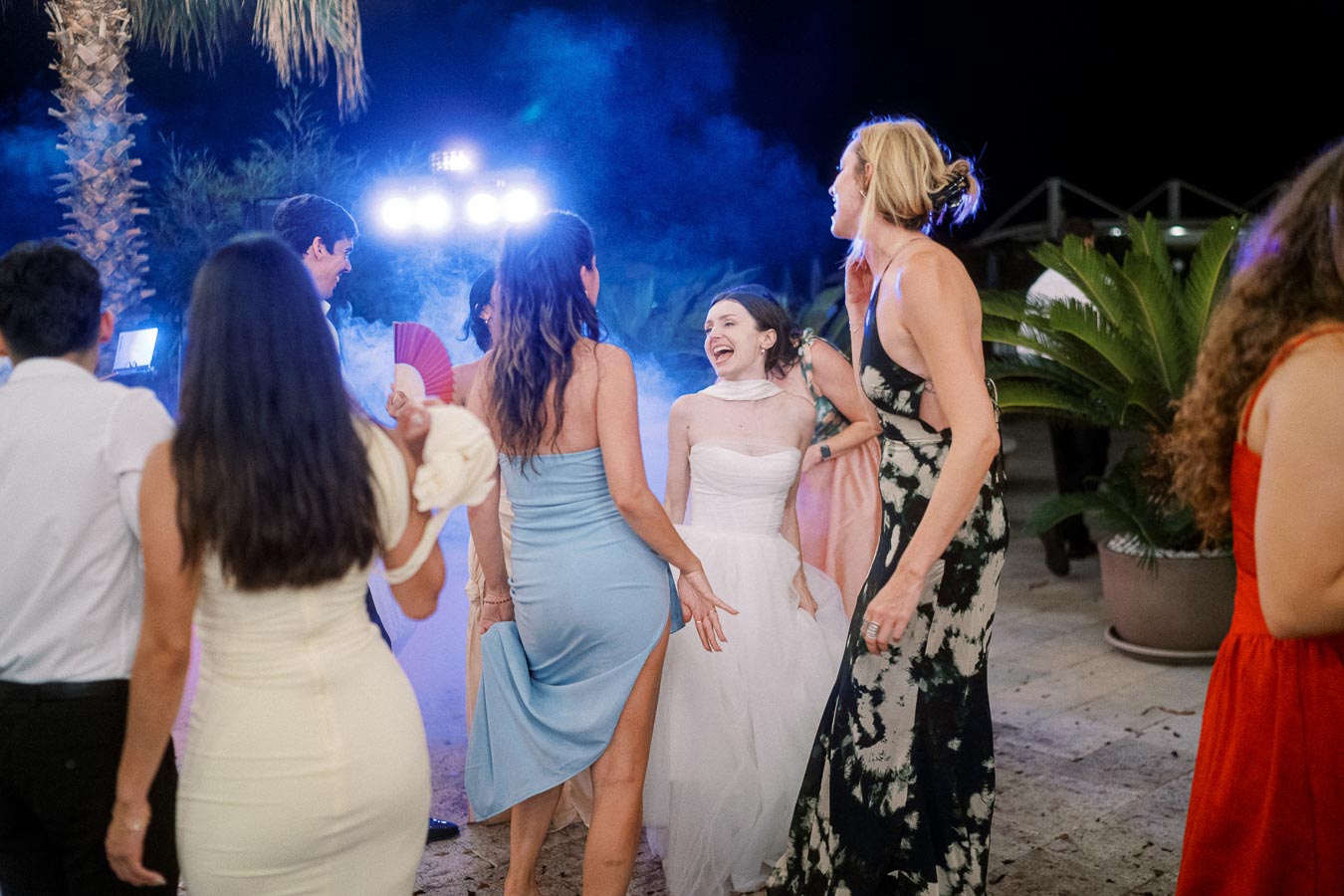 A lively wedding reception party with a group of elegantly dressed guests dancing and laughing under outdoor lighting, surrounded by tropical plants.