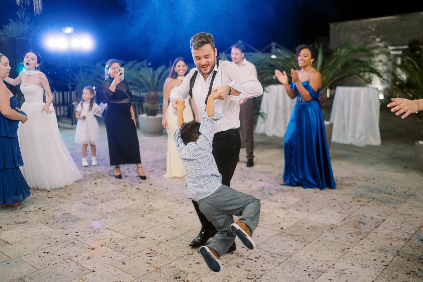 Man joyfully dancing with a child at an outdoor wedding reception, surrounded by elegantly dressed guests and blue atmospheric lighting.