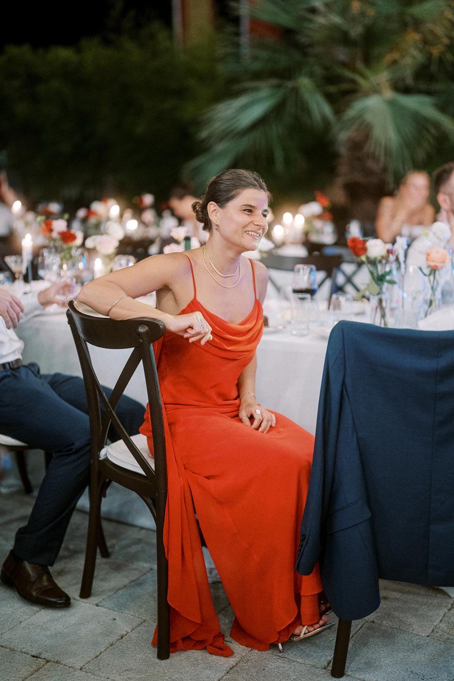 Smiling woman in elegant red dress sitting at an outdoor evening event with candlelit tables and floral decorations.