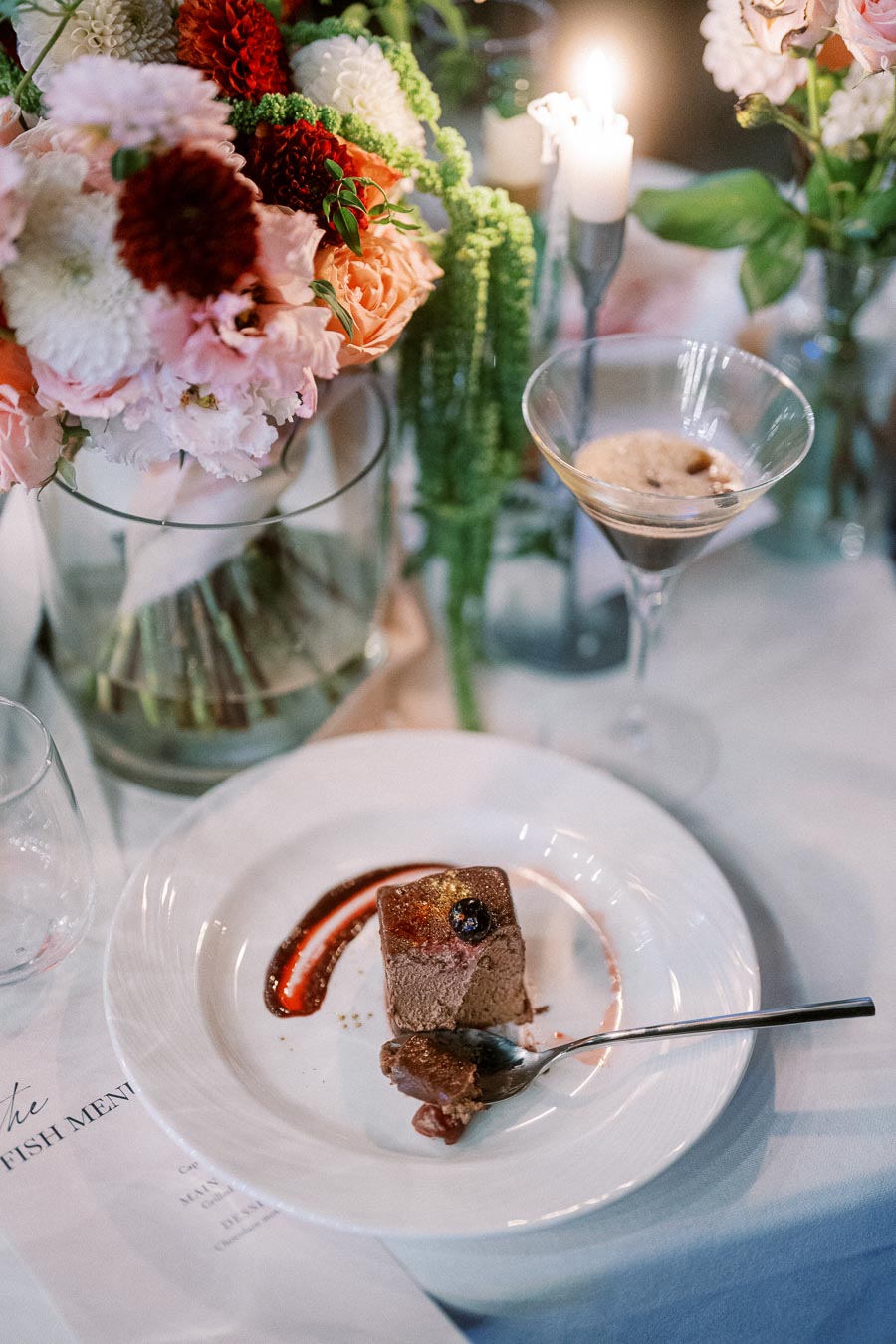 Elegant wedding table setting featuring a richly decorated centerpiece of vibrant flowers, accompanied by a gourmet dessert plated with red sauce and a drink in a martini glass, creating a sophisticated dining atmosphere.