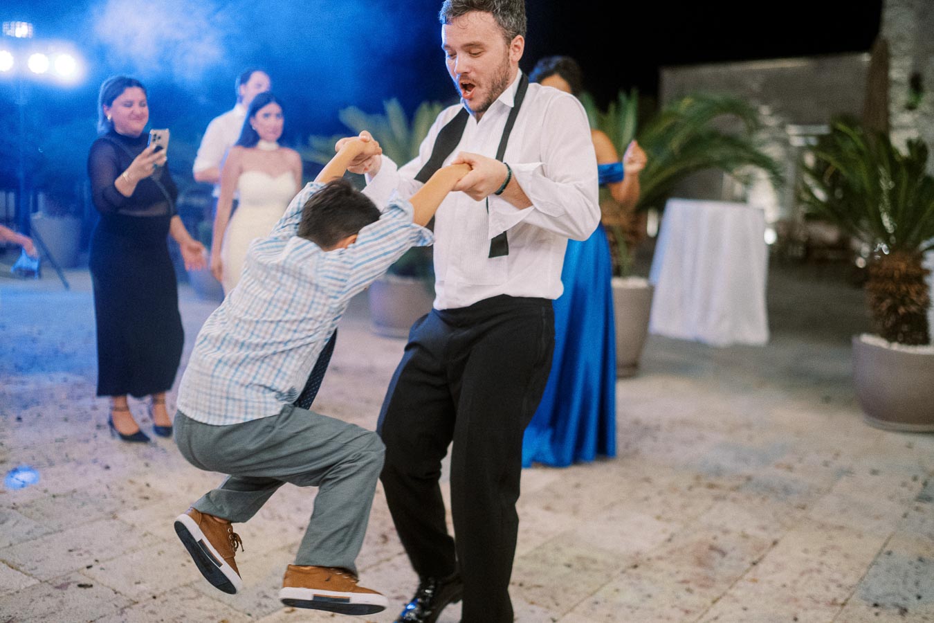 Man in formal attire joyfully dancing with a child at an outdoor evening event, surrounded by people in elegant dresses, capturing festive moments with a smartphone in the background.