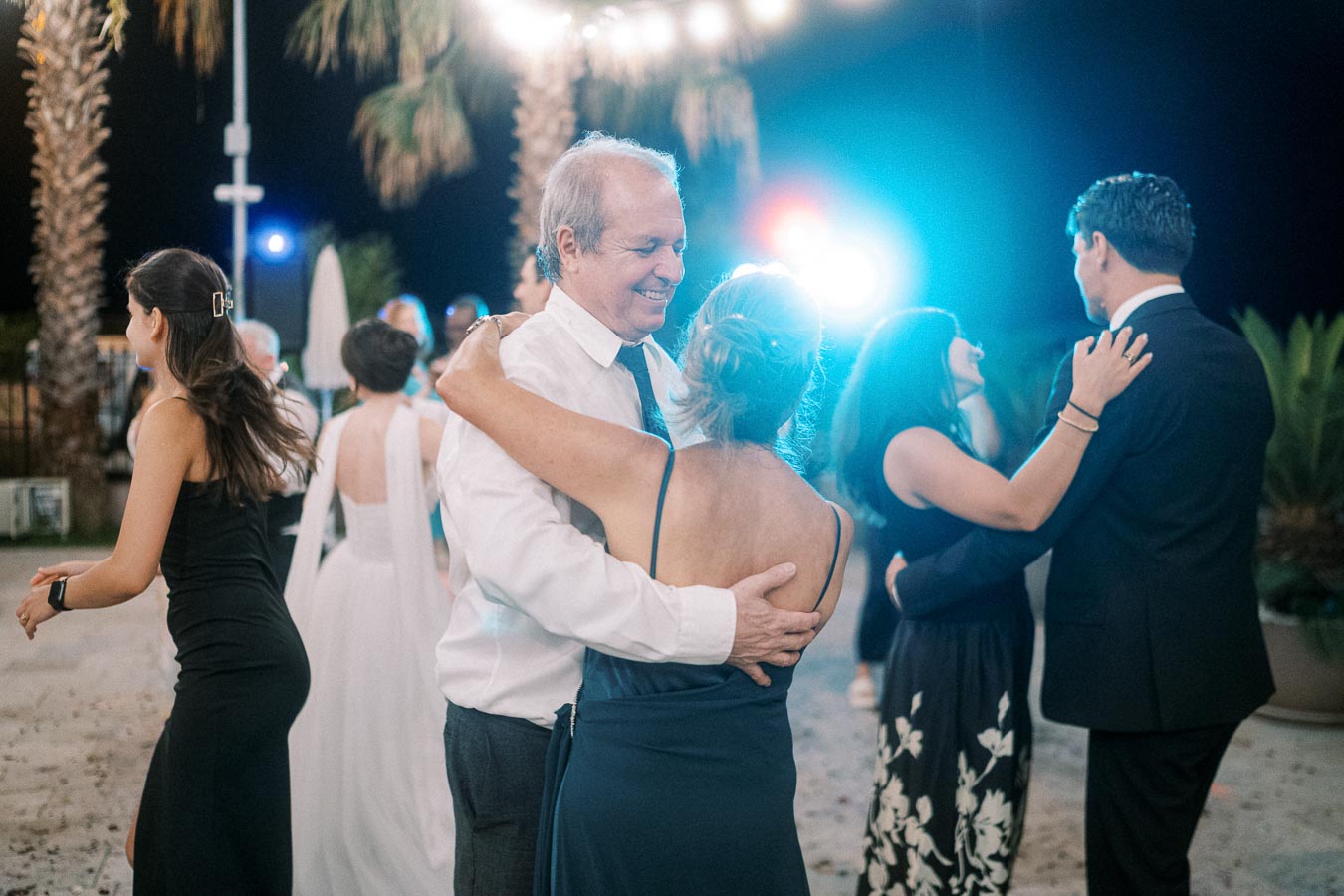 Couples dancing under string lights at an outdoor evening event, surrounded by palm trees, creating a festive and romantic atmosphere.