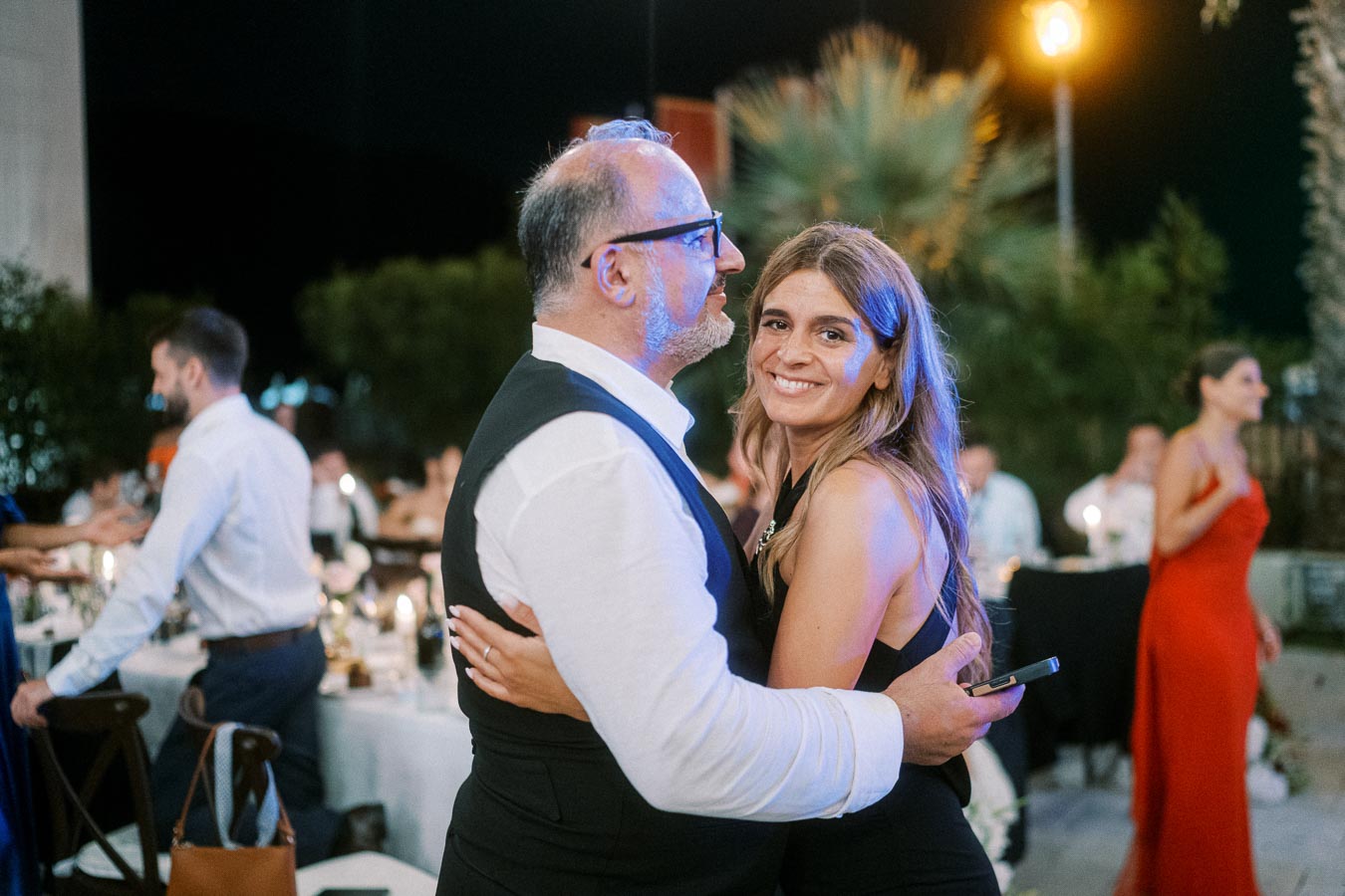 Couple embracing and smiling at an outdoor evening event, surrounded by elegantly dressed guests, with dim lighting and ambient decorations.