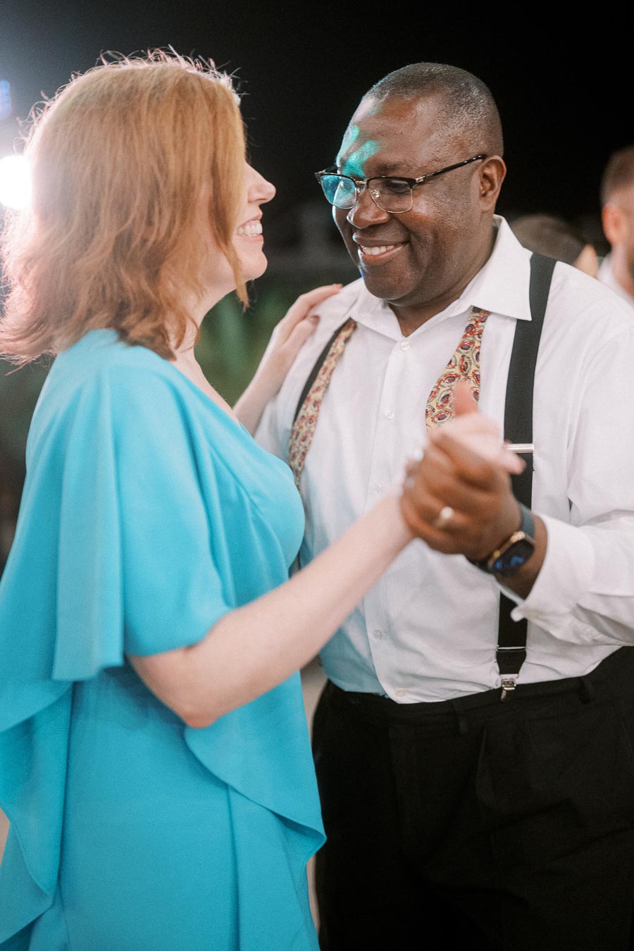 Couple joyfully dancing at an event, woman in blue dress and man in white shirt with patterned suspenders, sharing a happy moment.