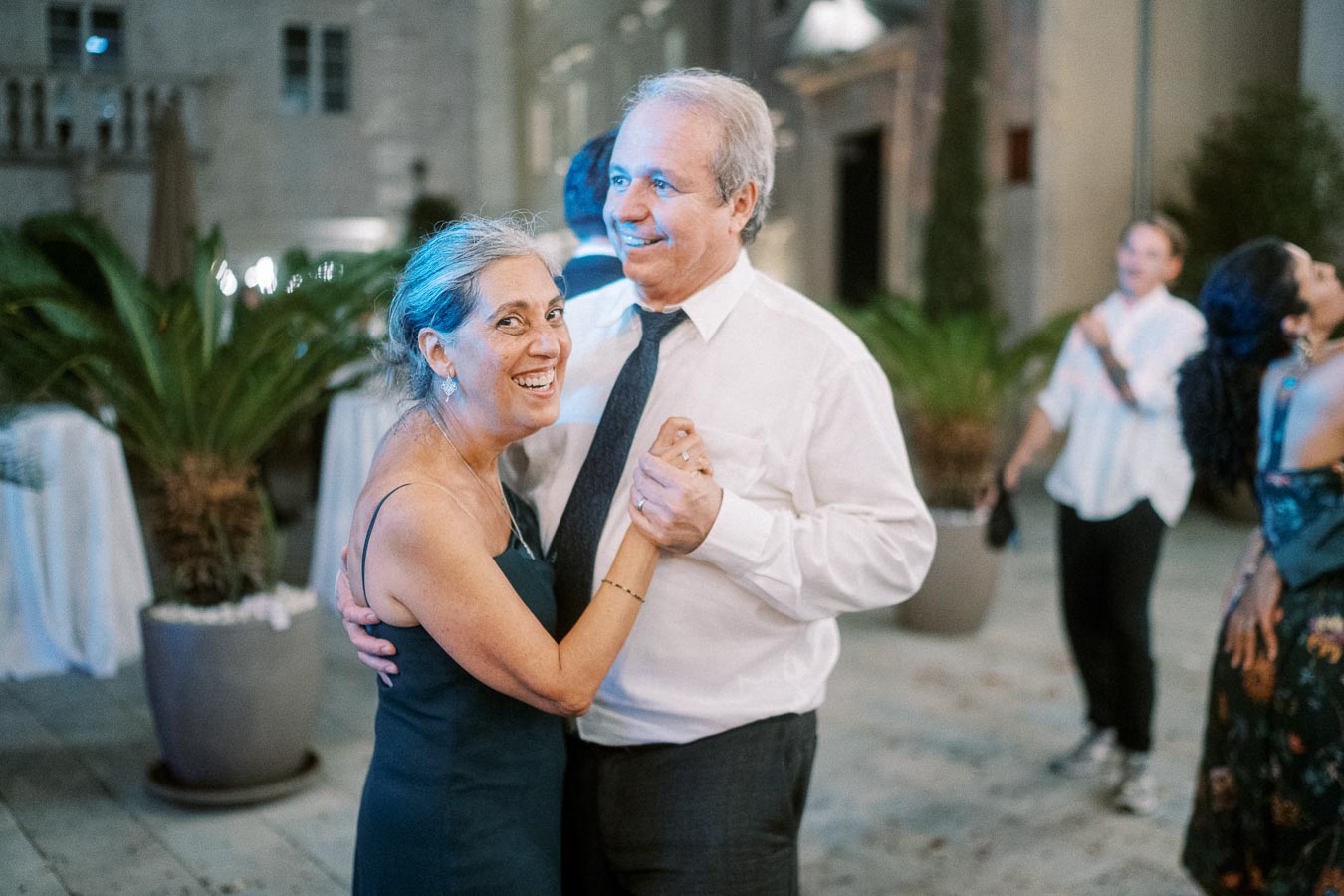 Elderly couple joyfully dancing at an outdoor evening event, surrounded by plants and other guests.