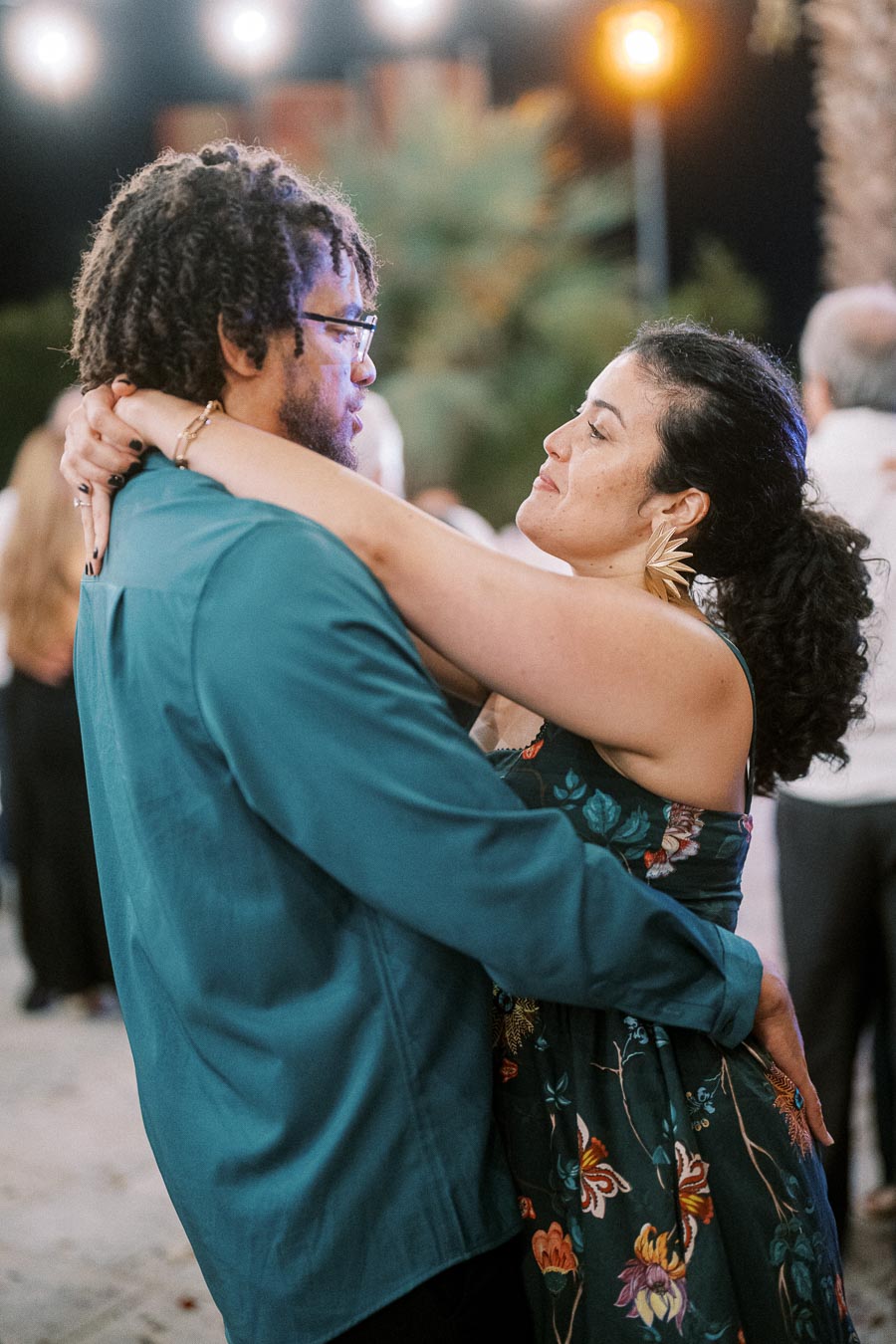 A couple dancing closely at an outdoor event, with festive lights in the background, creating a romantic atmosphere. The woman is wearing a floral dress and the man is in a teal shirt, both smiling at each other.