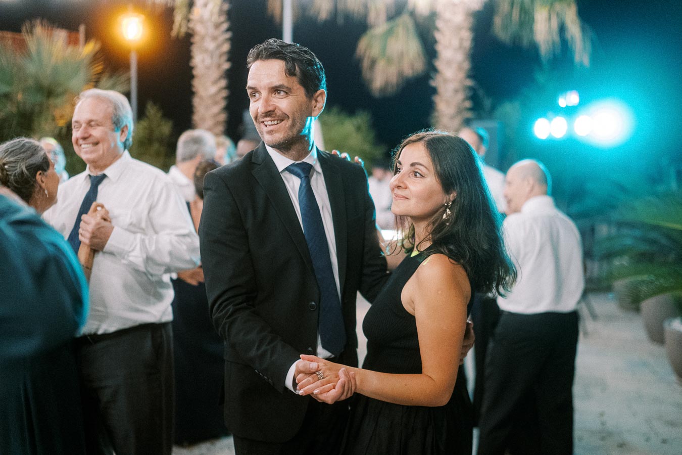 Couple enjoying a dance outdoors at a nighttime event, surrounded by other guests in formal attire, with palm trees and ambient lighting in the background.