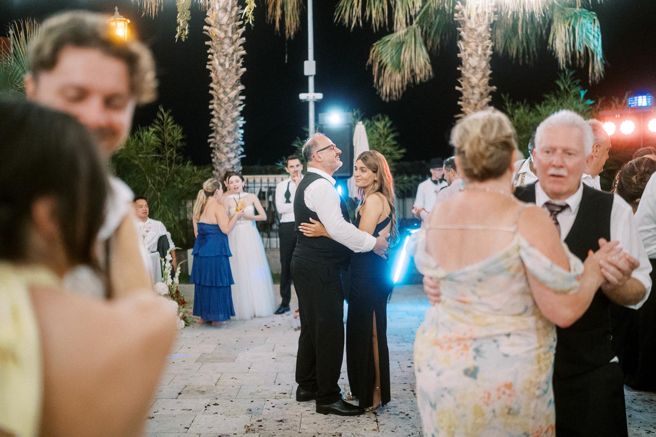 A lively outdoor wedding reception at night with couples dancing under palm trees, featuring elegant attire and festive lighting in a joyful atmosphere.