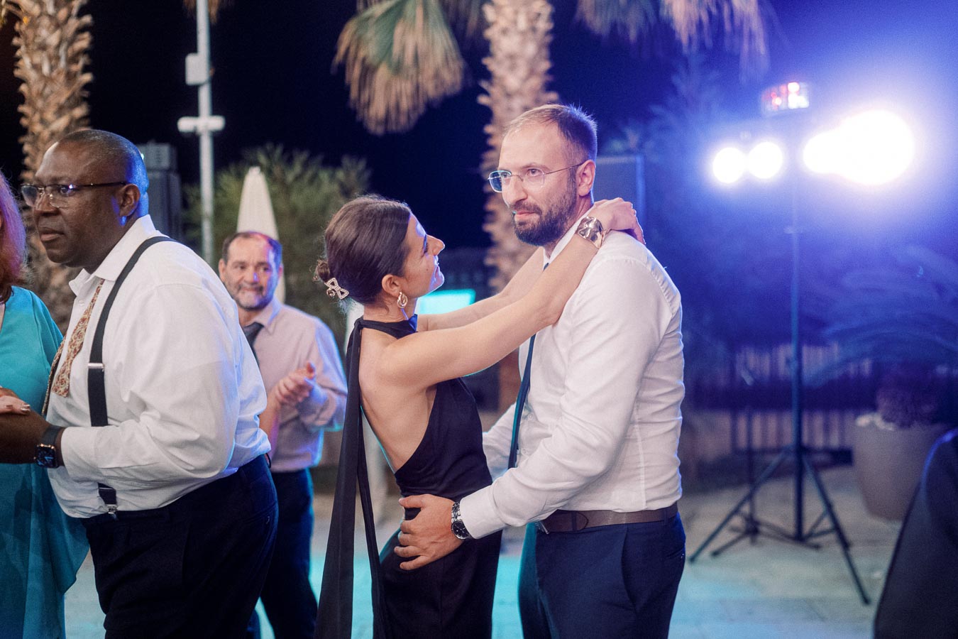 A couple dancing closely under evening lights at an outdoor event, surrounded by other attendees enjoying the festive atmosphere. Palm trees and soft lighting create a warm and romantic ambiance.