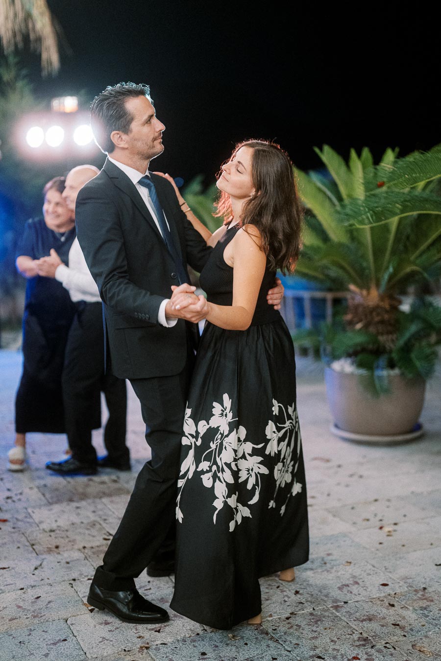 Elegant couple dancing at a nighttime outdoor event, with a man in a suit and woman in a floral black dress, surrounded by plants and soft lighting.