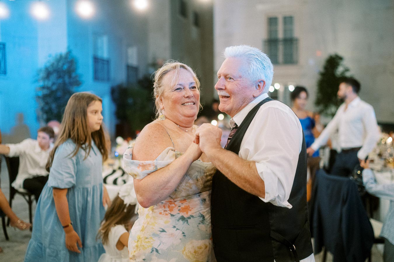Elderly couple joyfully dancing at a wedding reception, surrounded by guests in formal attire with ambient blue lighting.