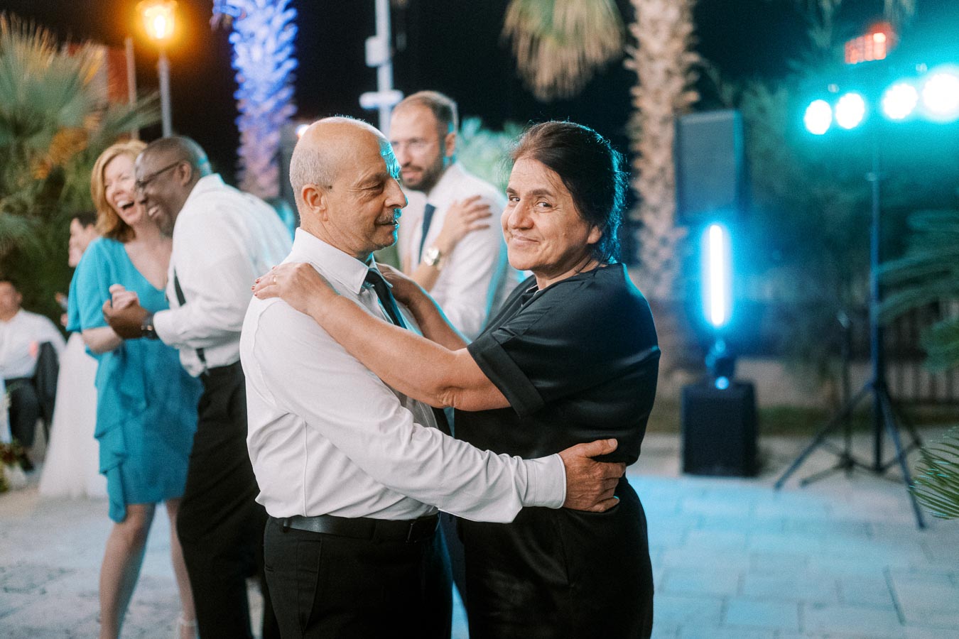 Elderly couple dancing together at an outdoor celebration, surrounded by joyful people, with decorative lights and palm trees in the background.