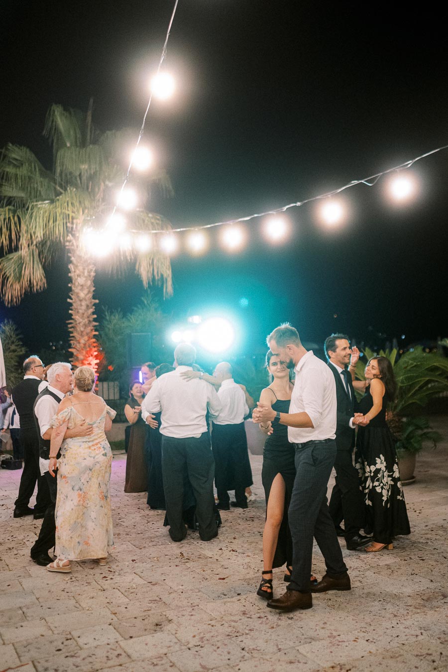 Couples dancing under string lights at an outdoor evening event with a tropical backdrop.