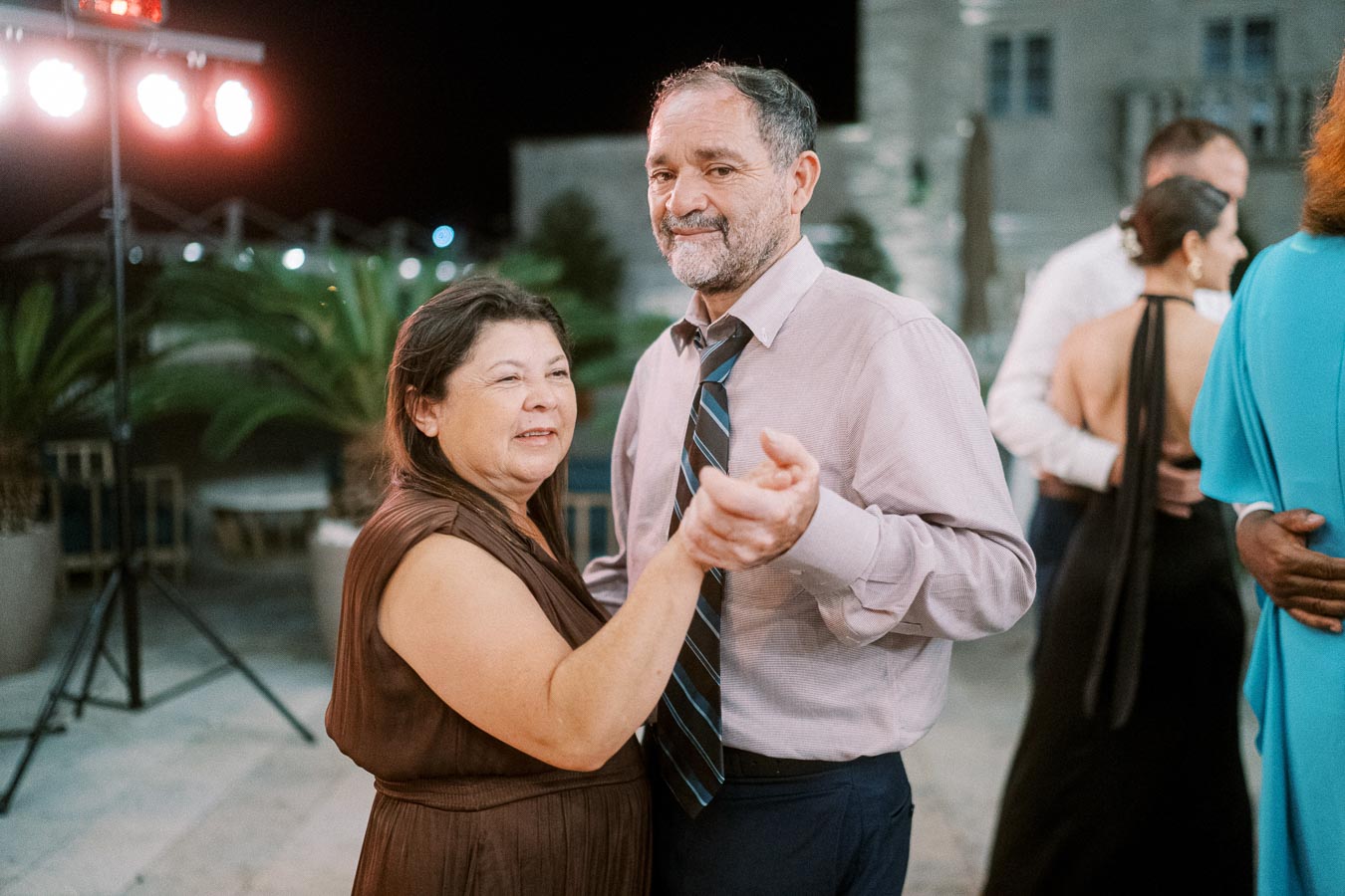 Elderly couple dancing at an outdoor evening event, with party lights and guests in the background.