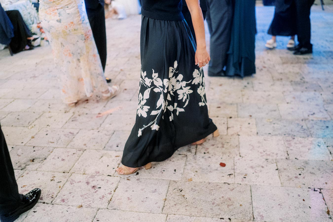 A person in a long, elegant black skirt with white floral patterns walks on a textured stone floor at a formal event.