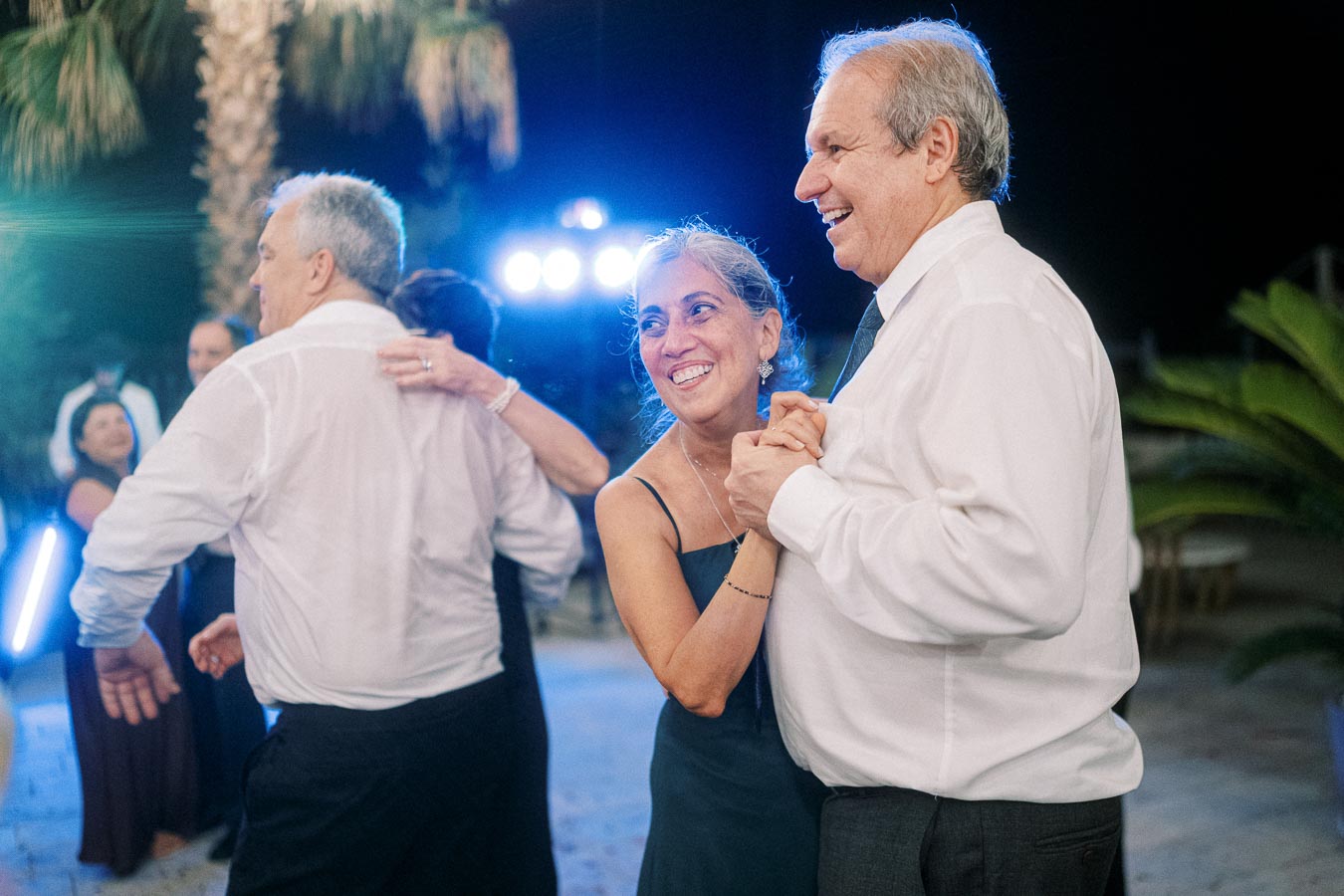Happy couple dancing at an outdoor evening event with palm trees in the background, illuminated by soft blue lights.