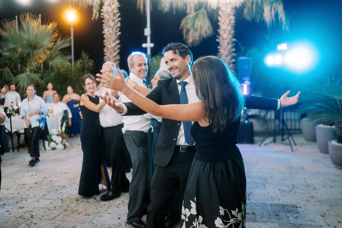 A lively outdoor celebration with people dancing joyfully under palm trees and string lights, creating a festive atmosphere at night.