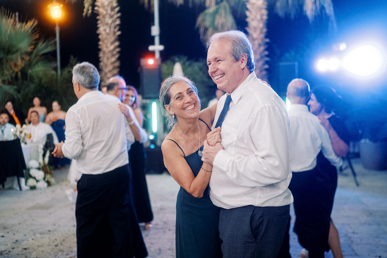 Elderly couple joyfully dancing under string lights at an outdoor evening event, surrounded by friends and tropical palm trees.