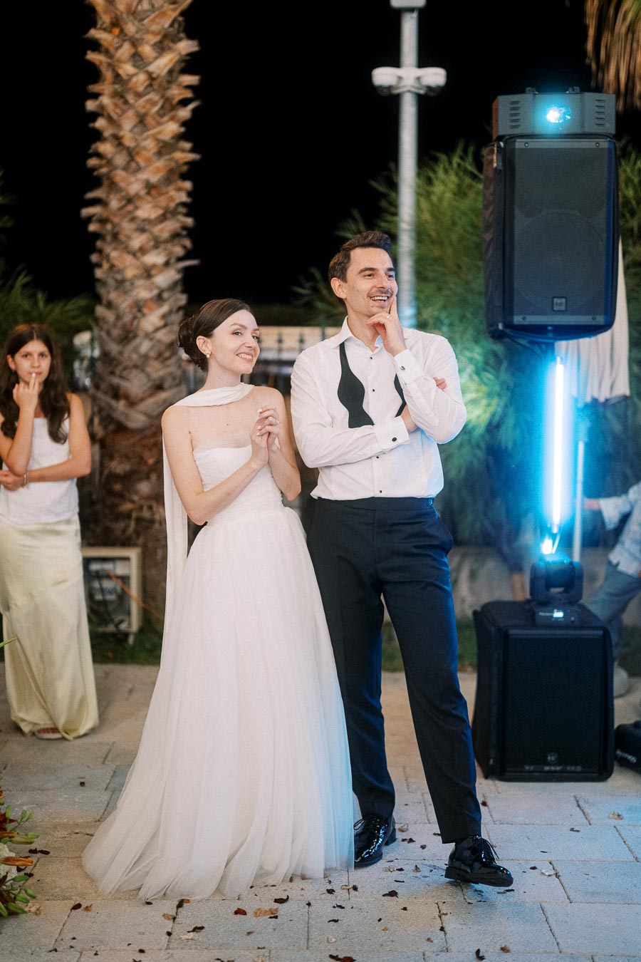 Bride and groom smiling and standing together at an outdoor evening wedding reception, with palm trees and a speaker in the background.