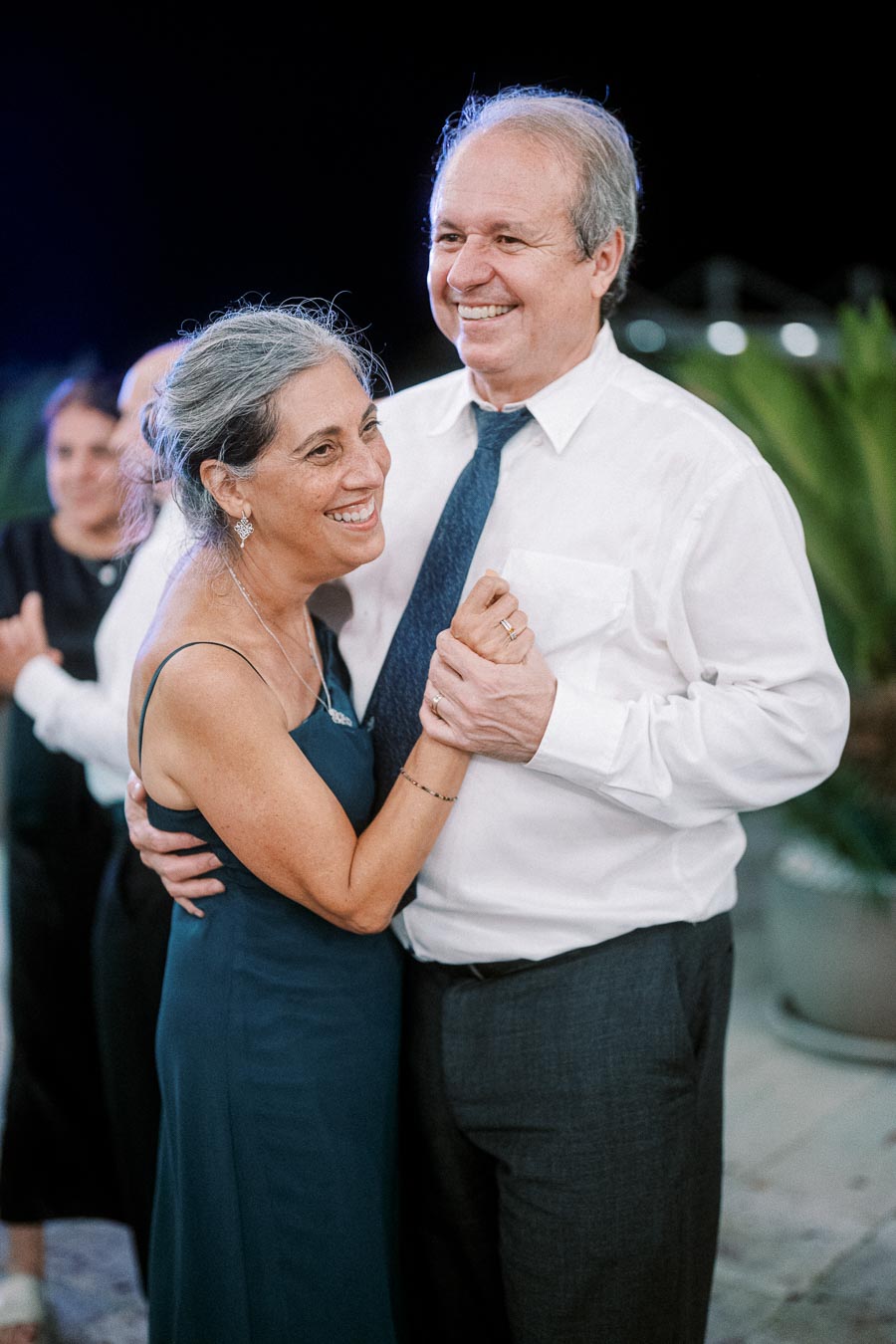 Elderly couple smiling and dancing together at a joyful celebration, dressed elegantly with the woman in a blue dress and the man in a white shirt and tie, surrounded by other guests in the background.