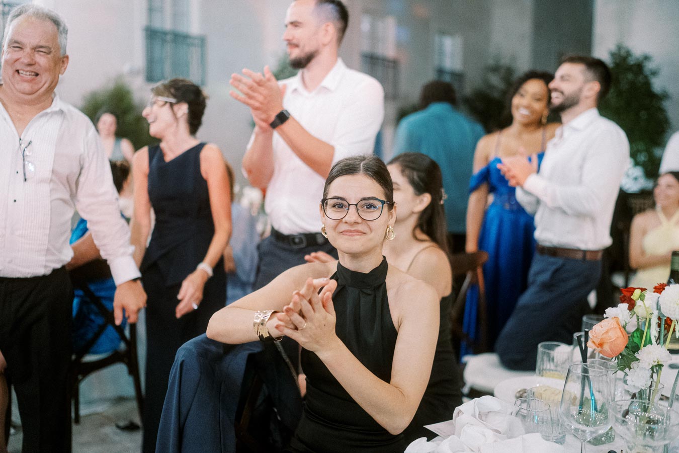 Group of people clapping and smiling at a formal event, with focus on a woman in glasses and a black dress seated at a table adorned with flowers and glasses.