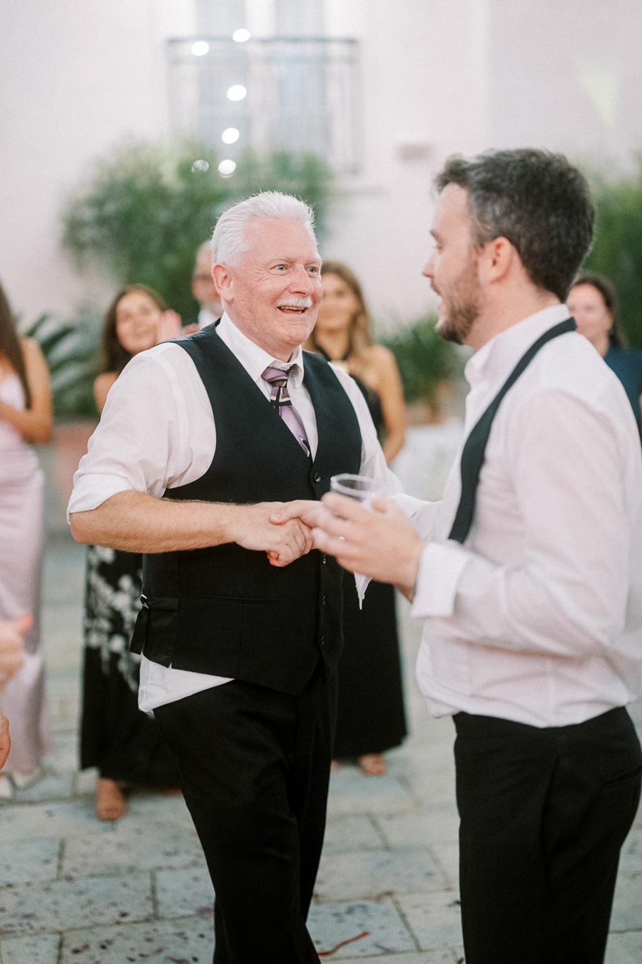 Senior man dancing joyfully with a younger man at an outdoor wedding reception, surrounded by smiling guests in formal attire.