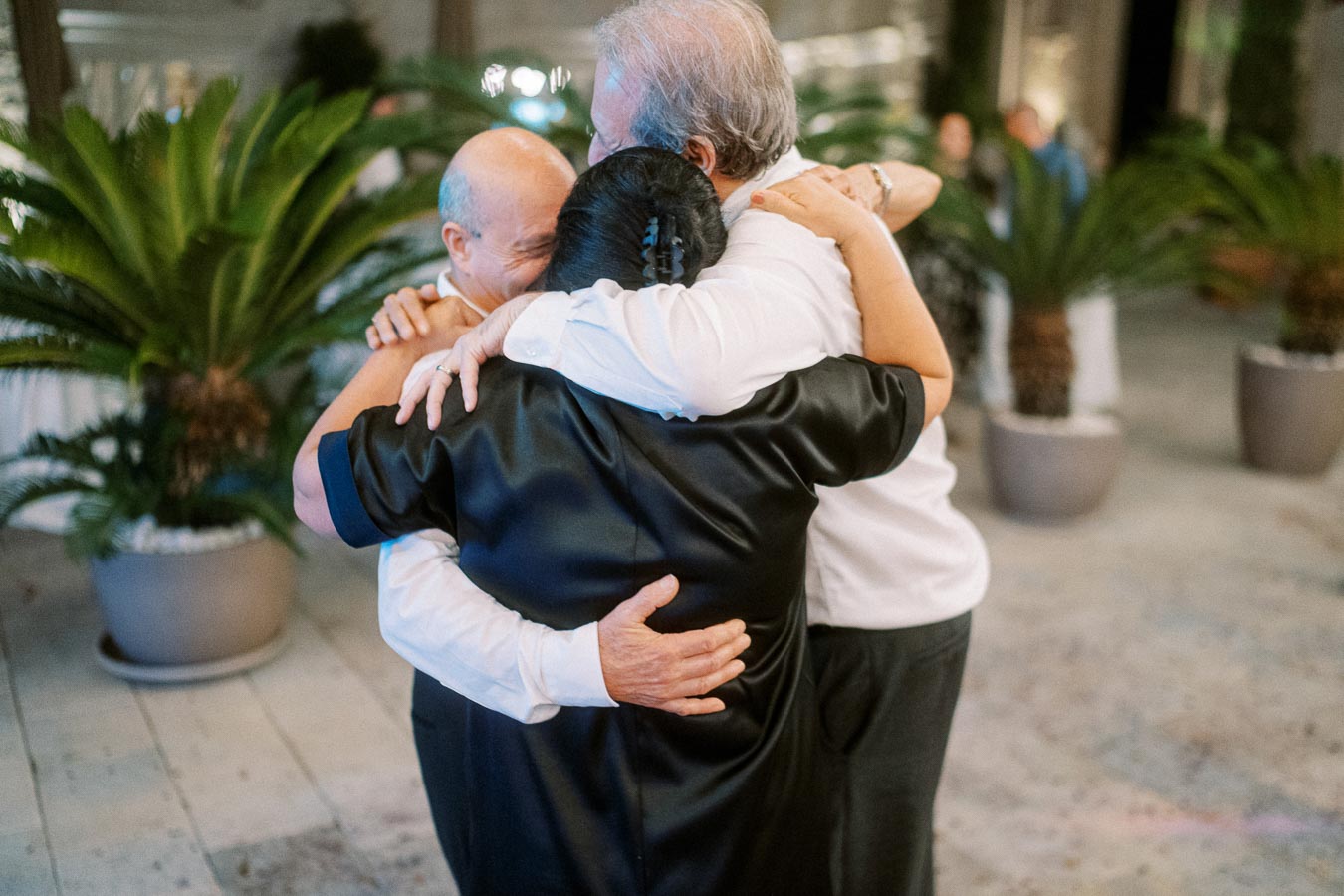 Three people embracing in a joyful group hug at an outdoor event with lush greenery in the background.