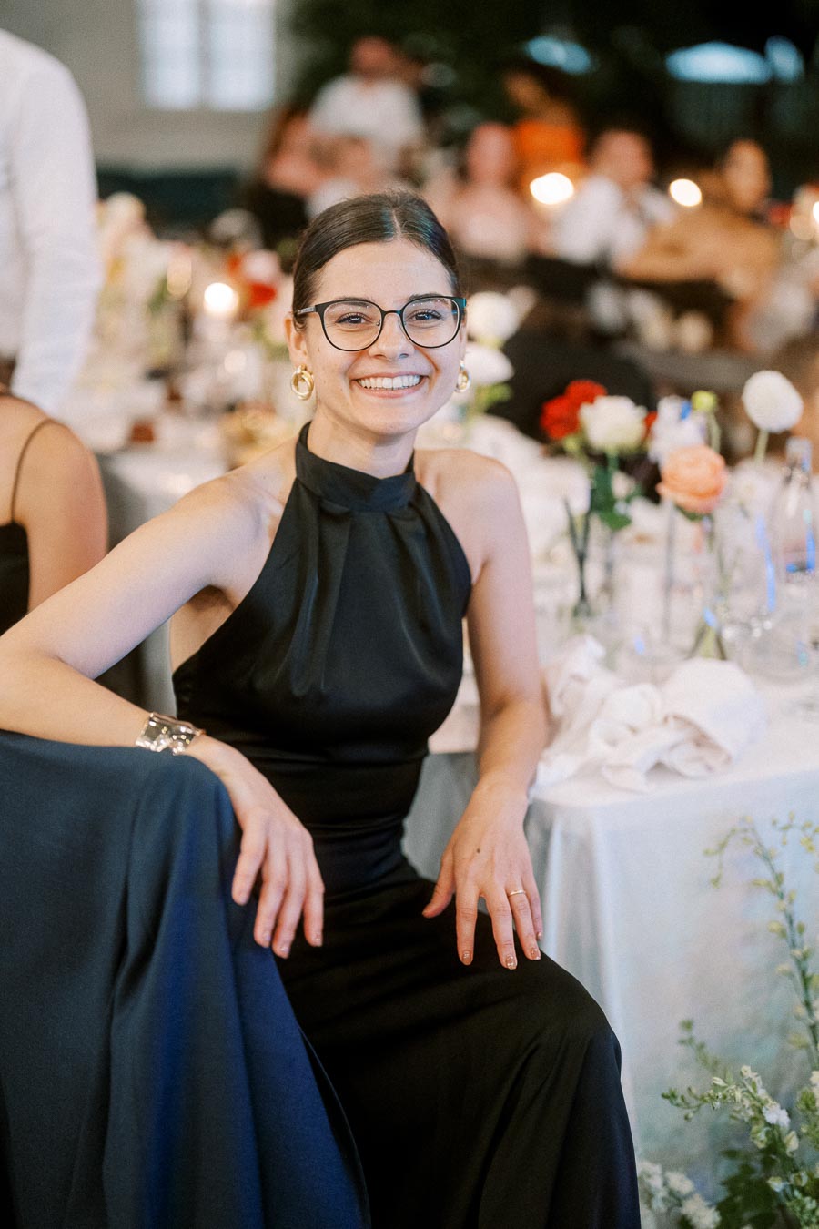 Smiling woman in black dress and glasses at a formal event, seated at a decorated table with flowers.